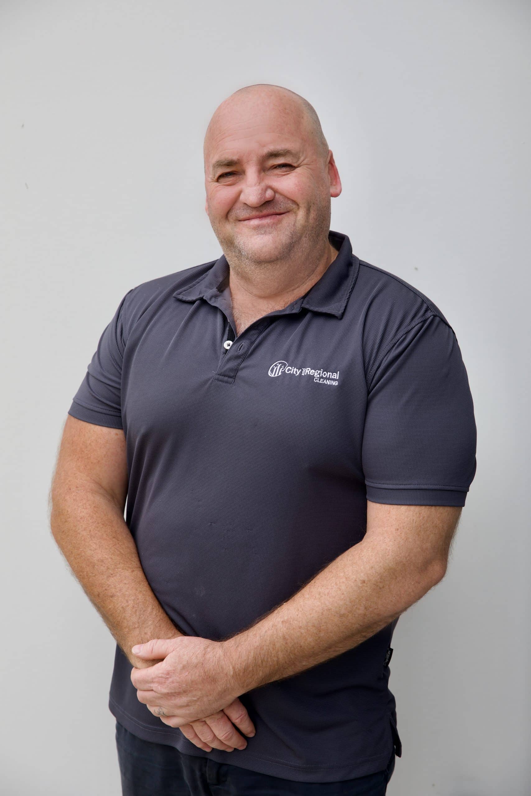 Man with a bald head smiles, wearing a dark polo shirt, arms crossed in front of a light background.