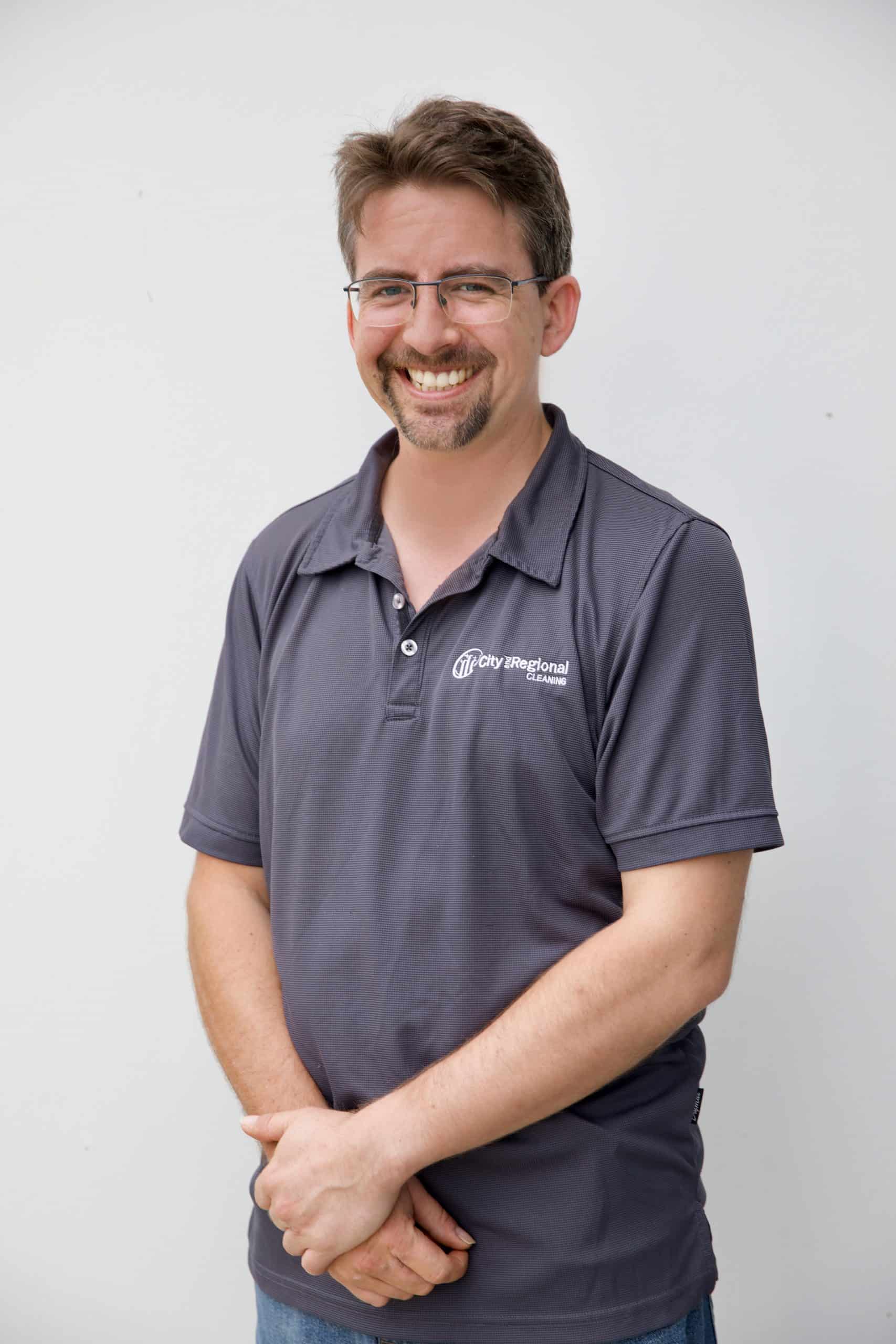Smiling man with glasses in a gray polo shirt, standing against a plain background.
