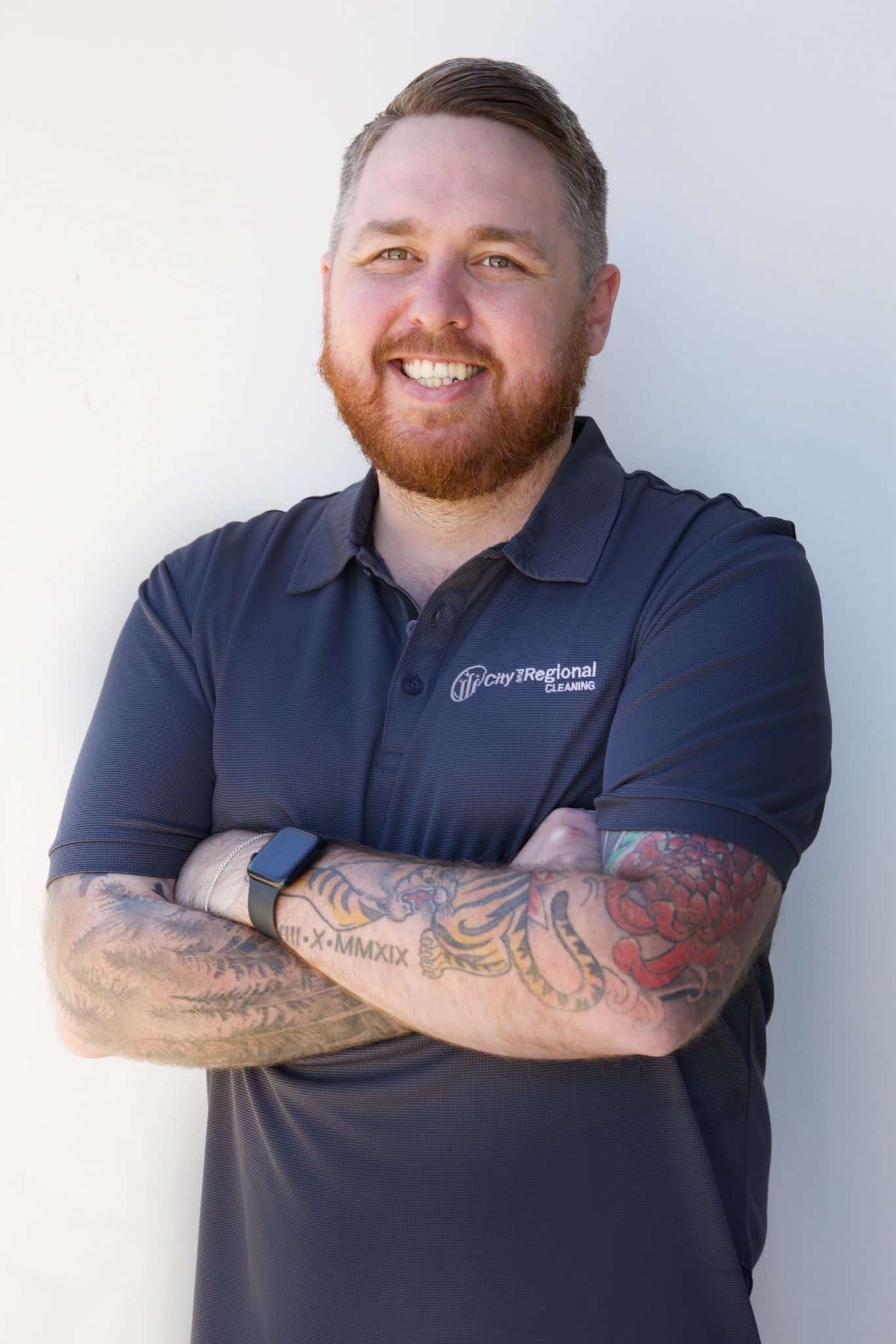 Smiling man with a beard, wearing a dark polo shirt, displays colorful arm tattoos.