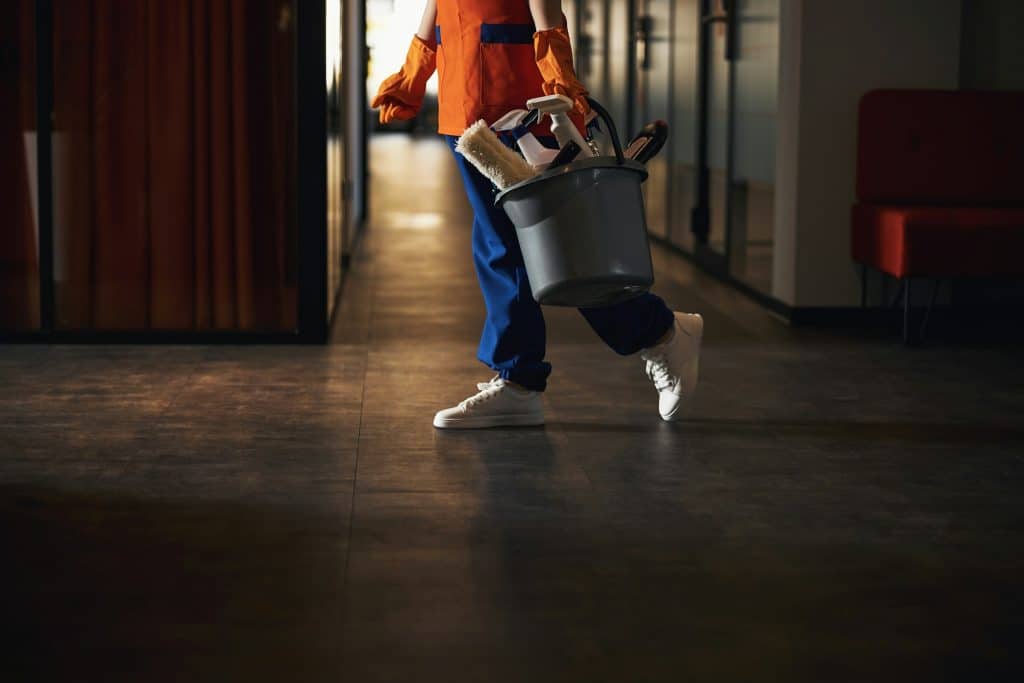 A person in orange and blue clothing carries a cleaning bucket down a hallway.