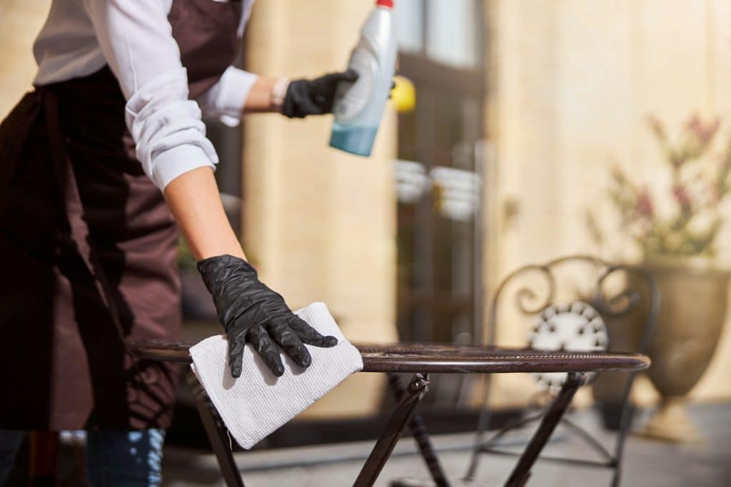 Person in a brown apron cleans a table with a cloth while holding a spray bottle.